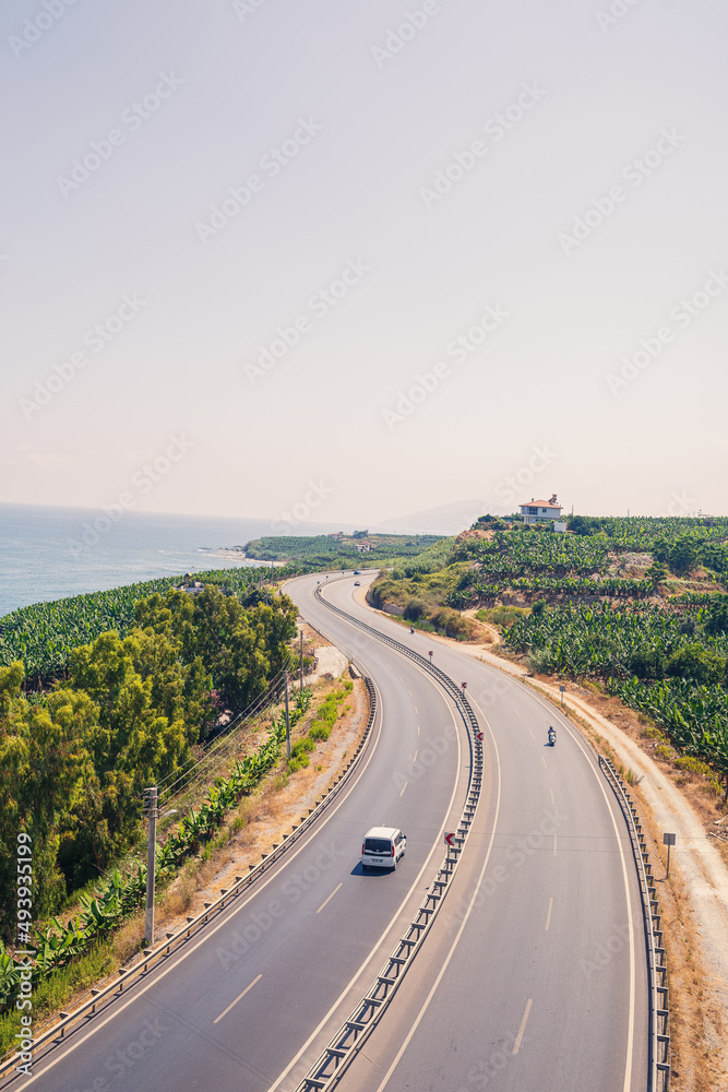 An empty asphalt road winds along the breathtaking scenic coastline on a sunny summer day. A spectacular shot of a coastal road overlooking clear blue skies and the calm Mediterranean Sea.