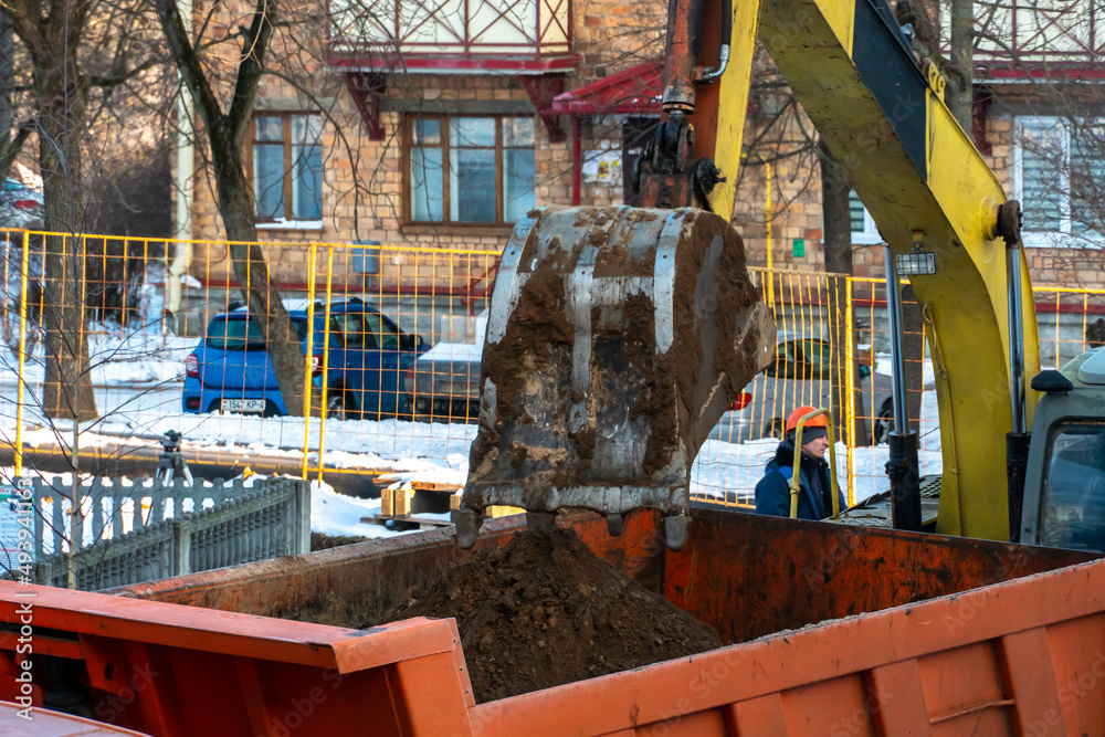 Naklejka premium Excavation works. The bucket of the tractor loads the soil with a shovel into the back of a dump truck on the construction site in winter. Earthworks with the help of heavy construction equipment