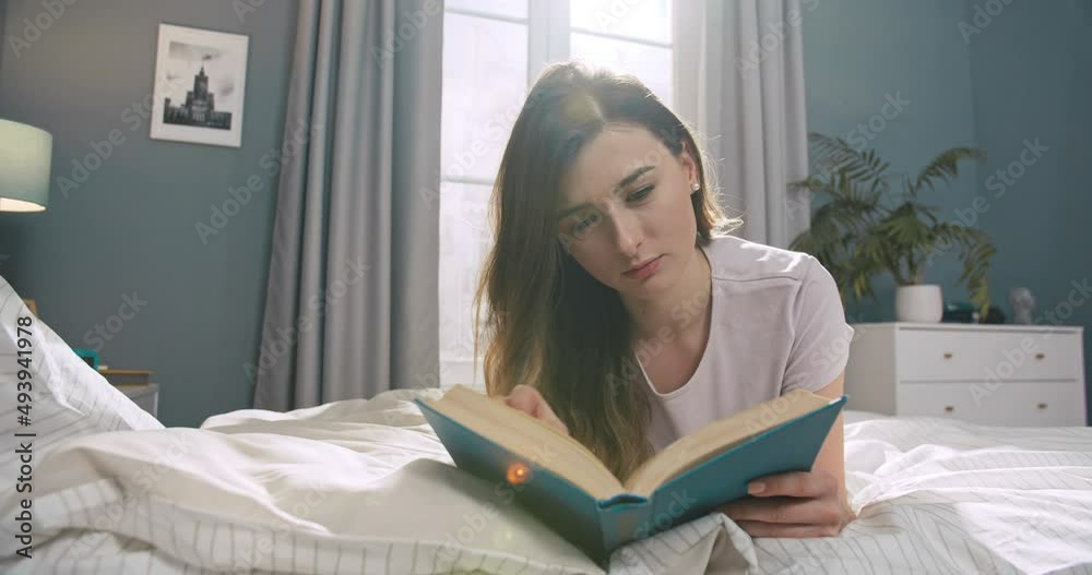 Young caucasian woman lying on the bed and reading a book early in the morning. Attractive woman enjoying her time lying on the bed in her apartment. Young caucasian woman lying on the bed and reading a book early in the morning. Attractive woman enjoying her time lying on the bed in her apartment.