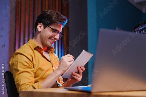 Young smiling professional fun content creator IT specialist programmer man in shirt work at office on laptop pc computer using tablet search information browse internet. Program development concept. photo