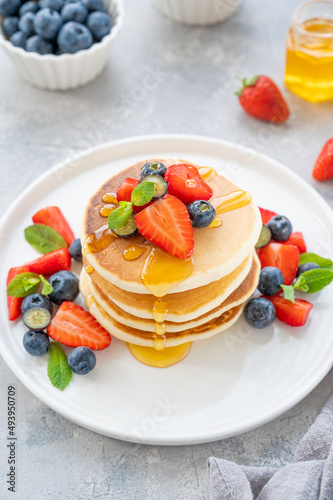 A stack of delicious fluffy pancakes with berries and honey on a white plate on a gray concrete background. Copy space.