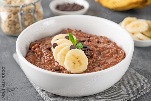 Chocolate oatmeal porridge with banana and chocolate chips on top in a white bowl. Healthy breakfast. Copy space.