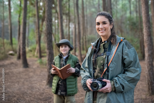 Woman with camera in forest. Female model in sportive clothes smiling at camera. Blurry boy in background. Hobby, photography concept