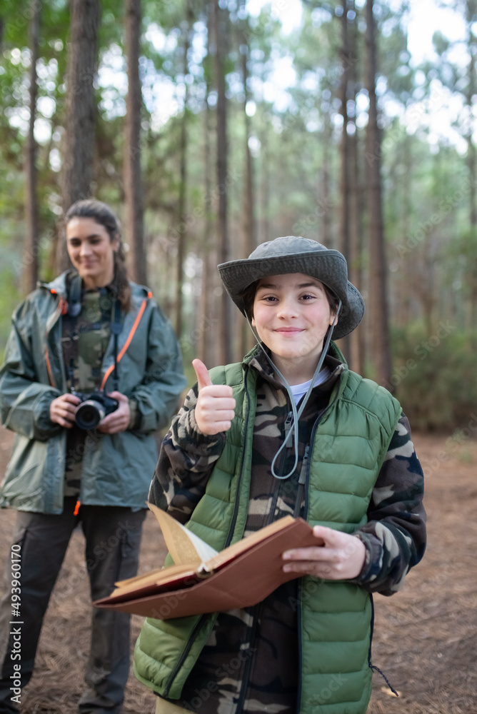 Fototapeta premium Joyful little boy with notebook in forest. Schoolboy in coat and panama smiling at camera, showing thumb up. Blurry mother in background. Childhood, nature, leisure concept