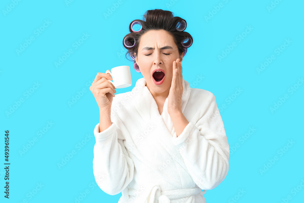 Sleepy young woman in bathrobe and hair curlers drinking coffee on blue background Sleepy young woman in bathrobe and hair curlers drinking coffee on blue background