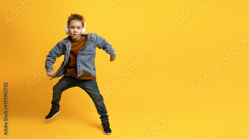full-length portrait of a 6-7-year-old kid in a denim shirt and a brown T-shirt with a hat on his head dancing with his arms outstretched, standing out against the pastel yellow background of child