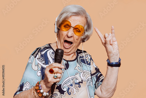 Funny grandmother portrait doing horns sign gesture with hand while singing karaoke using microphone and headphones at studio. Senior old woman dressing elegant for a special event. Rockstar granny.