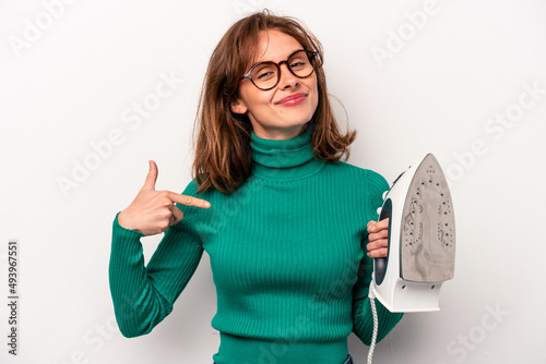 Young caucasian woman holding iron isolated on white background person pointing by hand to a shirt copy space  proud and confident