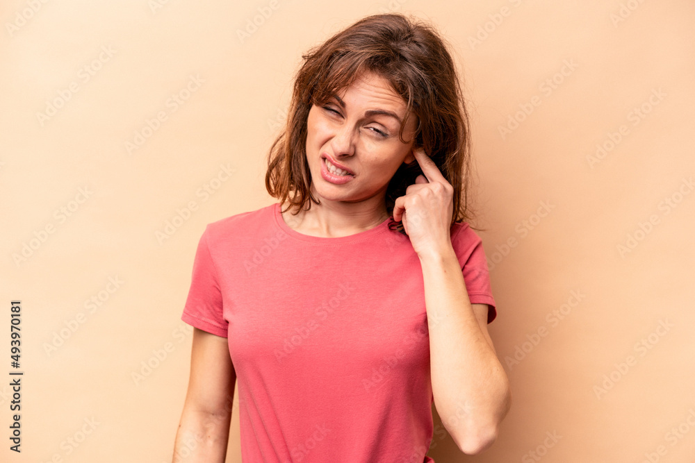 Young caucasian woman isolated on beige background covering ears with fingers, stressed and desperate by a loudly ambient. Young caucasian woman isolated on beige background covering ears with fingers, stressed and desperate by a loudly ambient.