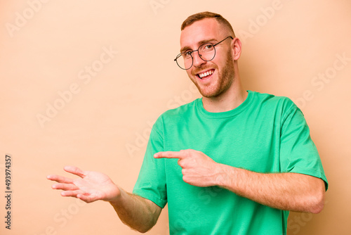 Young caucasian man isolated on beige background excited holding a copy space on palm.