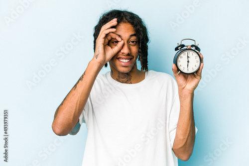 Young African american man holding alarm clock isolated on blue background excited keeping ok gesture on eye.