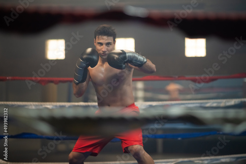 Young man in boxing gloves practicing kickboxing in ring. Through ropes view of concentrated sportsman in shorts training alone. Preparing for competition concept