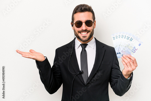 Young caucasian bodyguard man holding banknotes isolated on white background showing a copy space on a palm and holding another hand on waist. photo