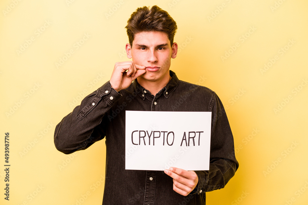 Young caucasian man holding crypto art placard isolated on yellow background with fingers on lips keeping a secret. Young caucasian man holding crypto art placard isolated on yellow background with fingers on lips keeping a secret.