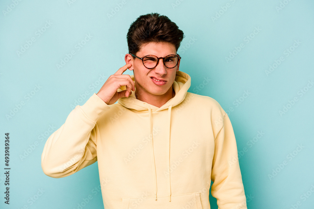 Young caucasian man isolated on blue background covering ears with fingers, stressed and desperate by a loudly ambient. Young caucasian man isolated on blue background covering ears with fingers, stressed and desperate by a loudly ambient.