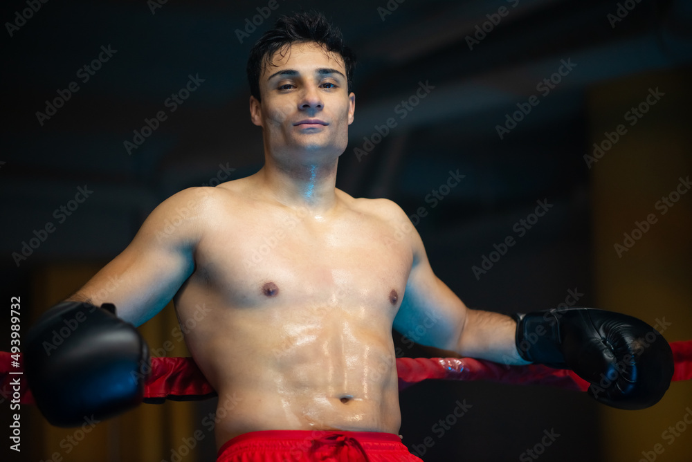 Confident boxer with sweaty torso leaning on rope. Portrait of serious young man in boxing gloves standing in ring, below view. Sportsman concept
