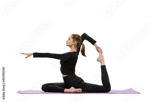 Portrait of young sportive woman in black tracksuit doing yoga and stretching exercises on purple mat isolated over white studio background