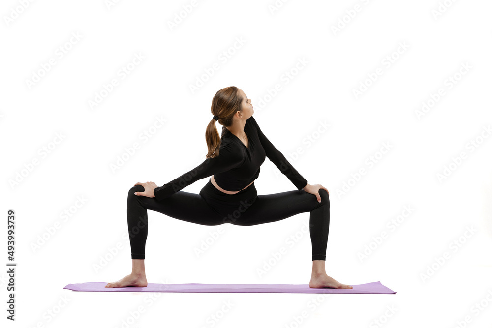Portrait of young gorl in black comfortable stracksuit training, doing stretching exercises on yoga mat isolated over white studio background