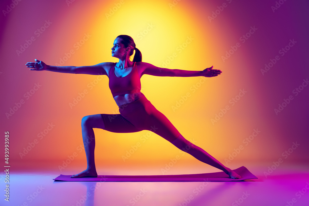 Portrait of young sportive girl doing yoga exercises isolated over gradient pink and yellow background in neon.