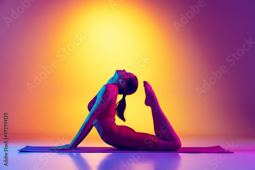 Portraity of young sportive girl doing stretching exercises, reaching head with legs isolated over gradient pink and yellow background in neon