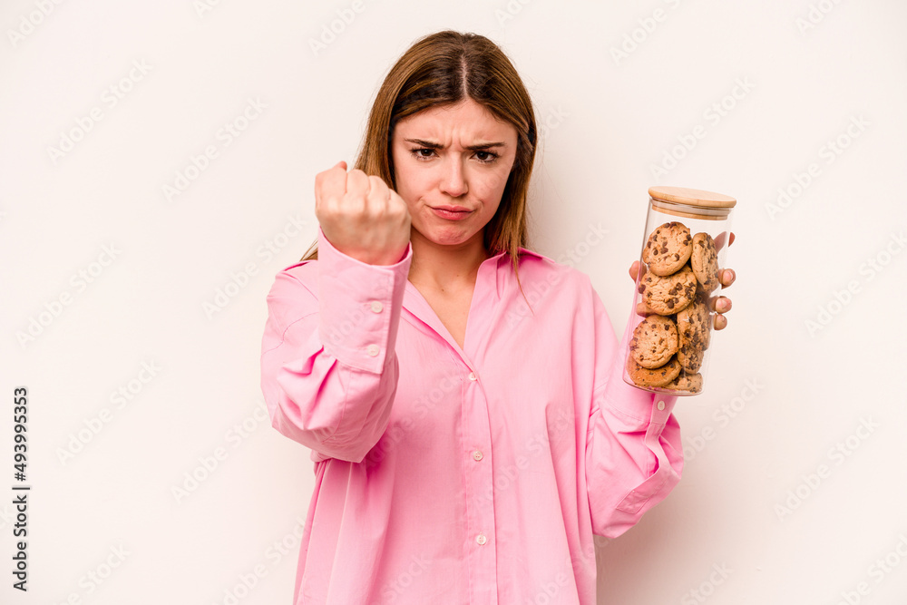 Young caucasian woman holding cookies jar isolated on white background showing fist to camera, aggressive facial expression.