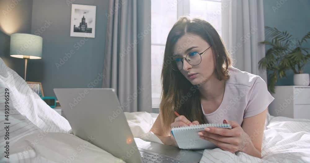 Young caucasian woman styding, making notes in paper notebook in bedroom. Pretty female wearing glasses, lying on bed and using laptop notebook to work. Young caucasian woman styding, making notes in paper notebook in bedroom. Pretty female wearing glasses, lying on bed and using laptop notebook to work.