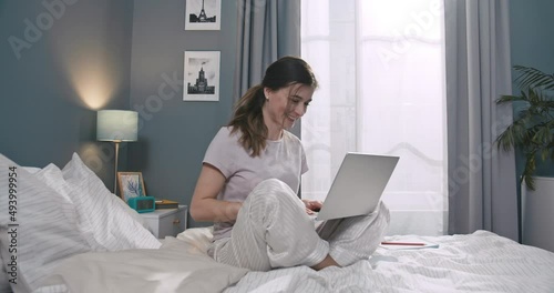 Beautiful caucasian woman sitting on bed, waving and making video call on computer. Young female spend leisure time at home, communicate on laptap in bedroom. photo