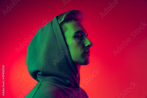 Portrait of young man in hood looking away, posing isolated over red studio background in green neon light