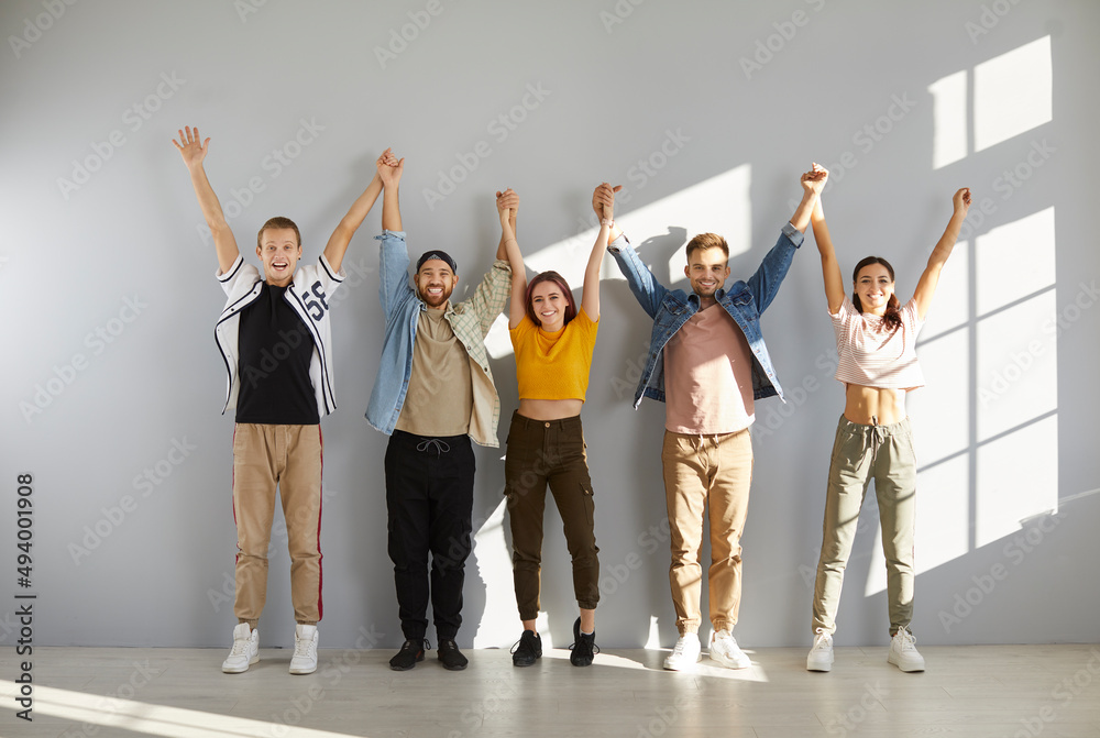 Obraz premium Happy people in casual clothes. Indoor studio group shot of young models in comfortable T shirts, denim jackets, pants, trousers, sneakers and trainers standing in row and holding and raising hands up