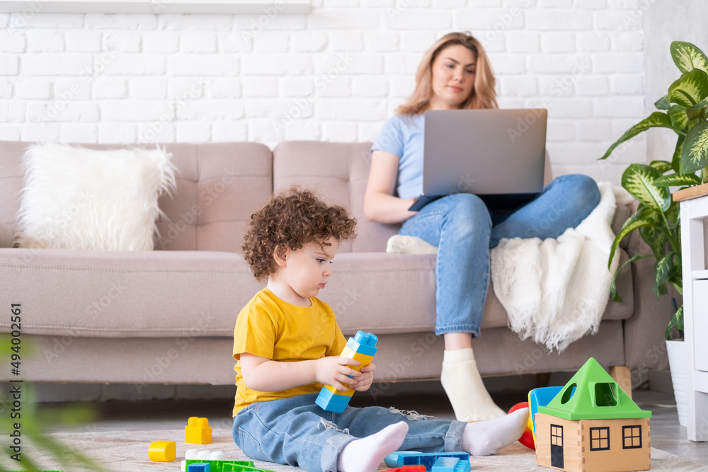 Mother freelancer using laptop and phone sitting on couch with kid daughter playing on smartphone Mother freelancer using laptop and phone sitting on couch with kid daughter playing on smartphone