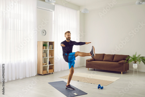Fit man having active workout at home. Handsome strong athlete following individual fitness program doing leg swing exercise on sports mat and using modern mobile phone app to track physical activity