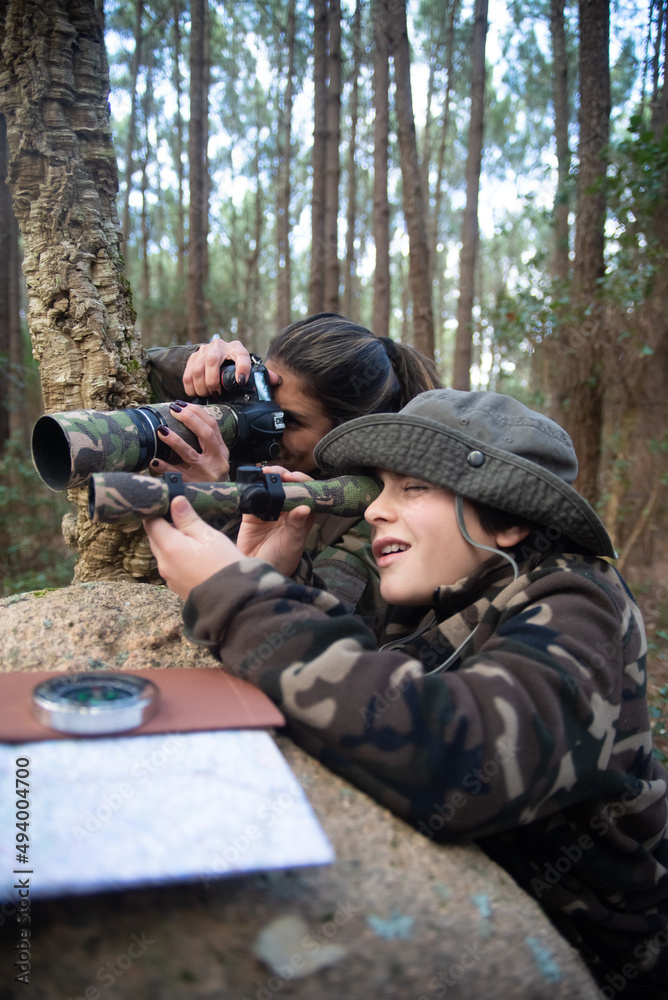 Fototapeta premium Pleased family taking pictures in forest. Mother and son with modern cameras lying on ground, peeking from stone. Parenting, family, leisure concept