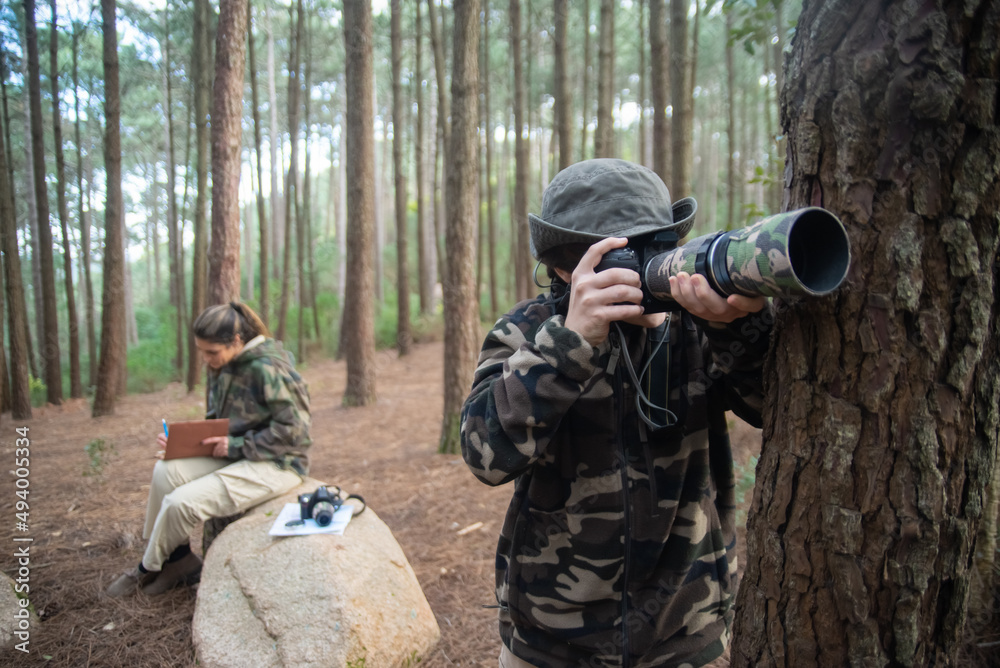 Fototapeta premium Absorbed boy with camera in forest. Schoolboy in coat and panama taking pictures. Blurry mother in background. Childhood, nature, leisure concept