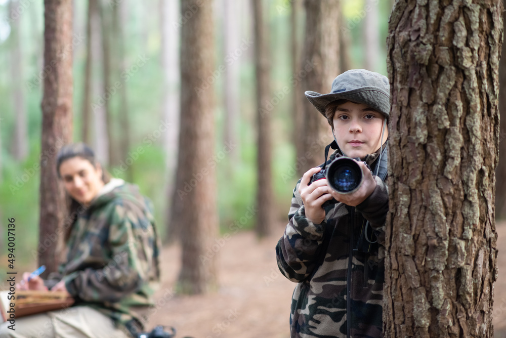Serious boy with camera in forest. Schoolboy in coat and panama looking at camera. Blurry mother in background. Childhood, nature, leisure concept Serious boy with camera in forest. Schoolboy in coat and panama looking at camera. Blurry mother in background. Childhood, nature, leisure concept