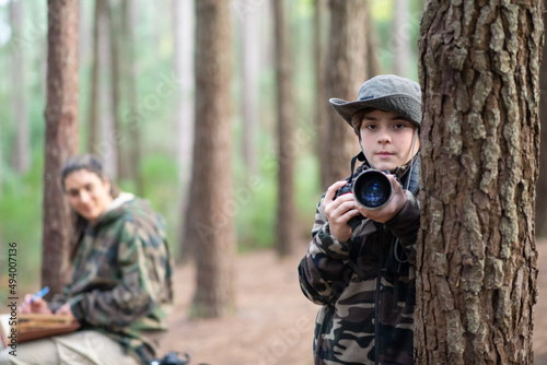 Serious boy with camera in forest. Schoolboy in coat and panama looking at camera. Blurry mother in background. Childhood, nature, leisure concept