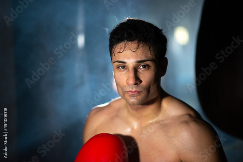 Brunette shirtless boxer in dark gym. Portrait of serious young Portuguese man with sweat on body standing against backlit. Dedication concept