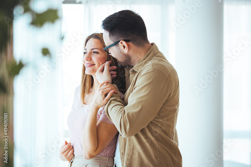 Loving couple embracing and sitting near window. Happy girlfriend and smiling boyfriend looking away and thinking about their future together. Young man and woman contemplating.