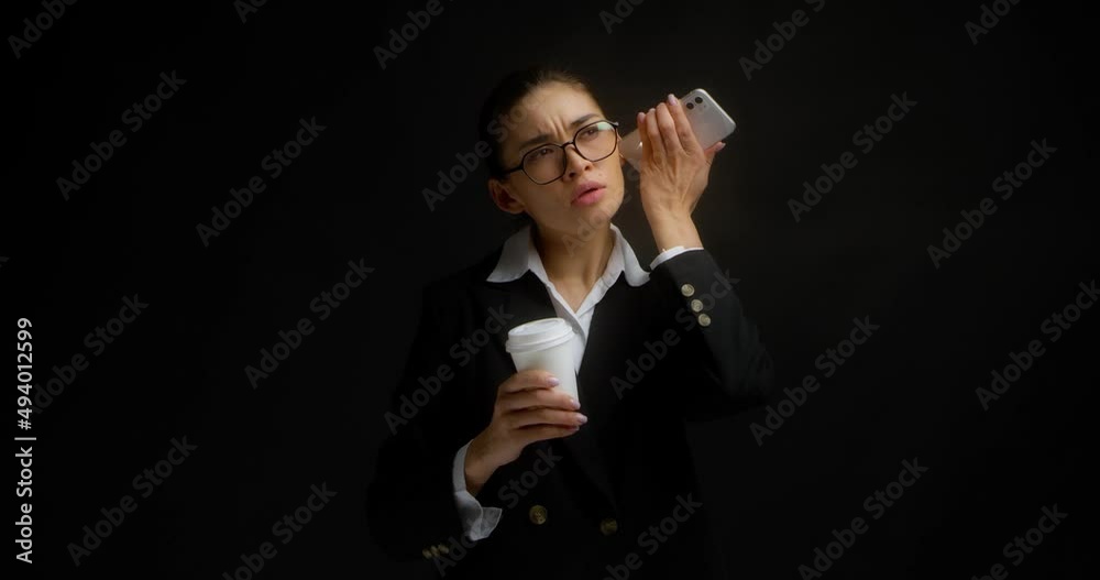 Business woman in glasses and black and white clothes is holding coffee in a white paper cup and listening to an audio message from a messenger on her cell phone. Isolated on a black background Business woman in glasses and black and white clothes is holding coffee in a white paper cup and listening to an audio message from a messenger on her cell phone. Isolated on a black background