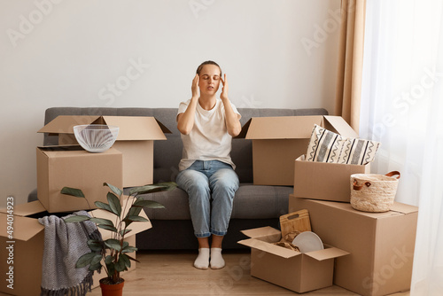 Image of attractive tired woman wearing white t shirt and jeans sitting on sofa during relocating to a new apartment, being exhausted and trying to concentrate. photo