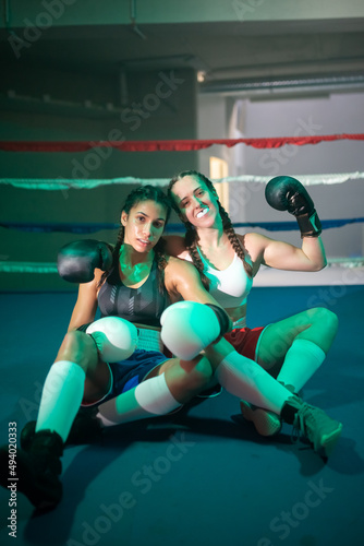 Portrait of happy boxer girls sitting on ring floor. Two beautiful girls in sportswear and gloves resting after active sparring, hugging looking at camera. Healthy lifestyle and combat sport concept