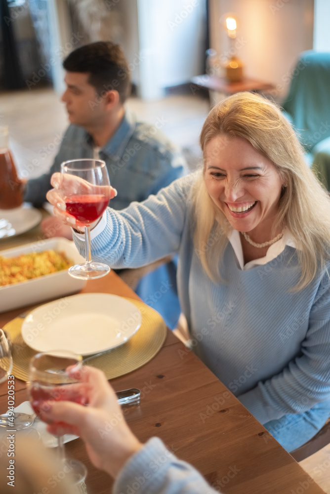 Cheerful mid adult woman raising wine glass at dinner party. Family having meal at home together. Family party concept Cheerful mid adult woman raising wine glass at dinner party. Family having meal at home together. Family party concept
