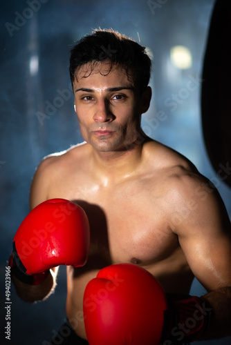Young Portuguese boxer without shirt standing in dark, smoky gym. Portrait of serious muscular man in boxing gloves against light. Fighter concept