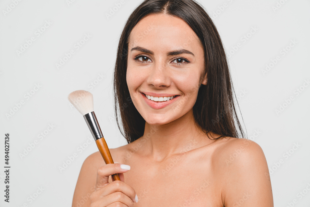 Caucasian young attractive woman girl lady with bare shoulders applying makeup with cosmetics brush on her face, doing daily care isolated in white background Caucasian young attractive woman girl lady with bare shoulders applying makeup with cosmetics brush on her face, doing daily care isolated in white background