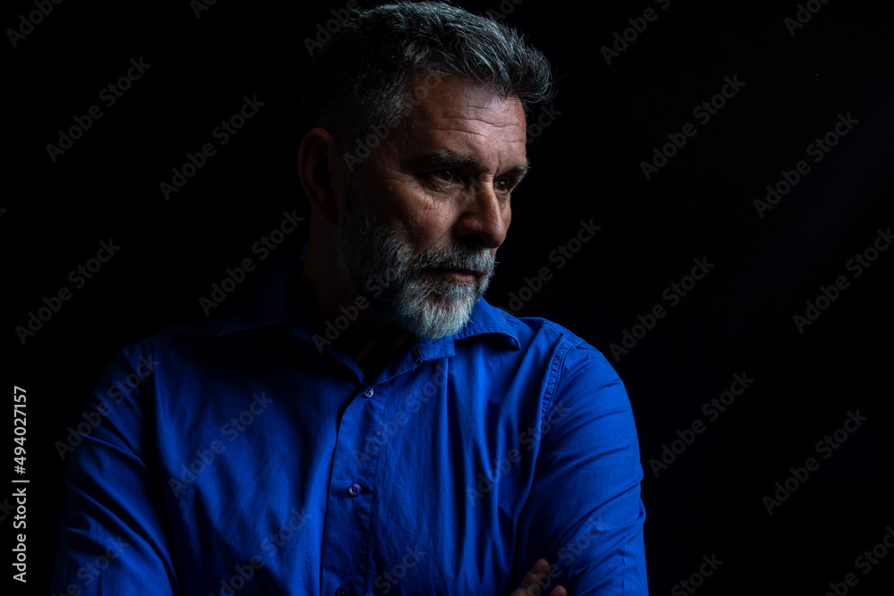 Dramatic portrait of a man in a low key. Studio low key portrait of a mature bearded man wearing a blue shirt. Portrait of an attractive bearded man in low key studio.