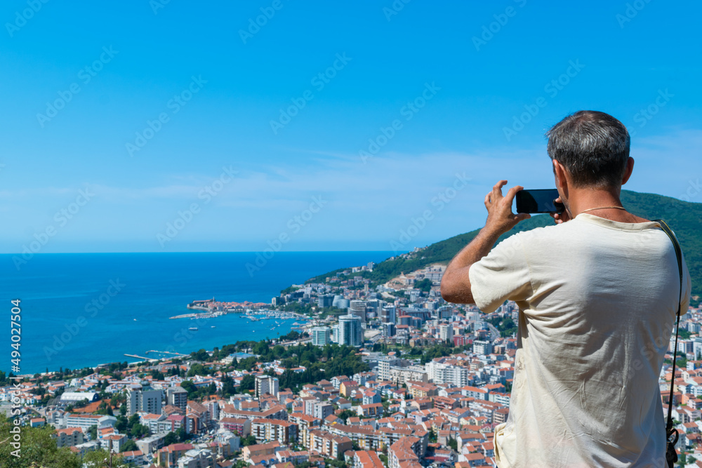 Man tourist takes a photo of the architecture Budva city from a hilltop. Panoramic landscape. Top view. Adriatic sea. Montenegro Man tourist takes a photo of the architecture Budva city from a hilltop. Panoramic landscape. Top view. Adriatic sea. Montenegro