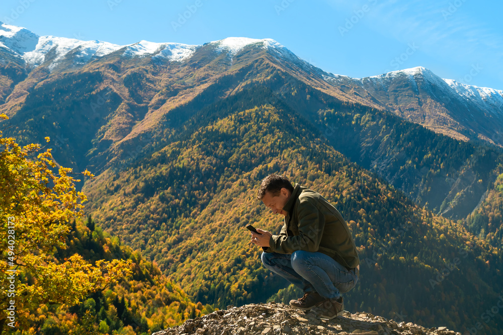 Young male tourist blogger is sitting on a slope with a mobile phone in his hands against the background of mountains on an autumn sunny day