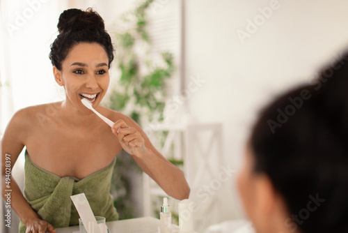 Lovely young lady in towel brushing teeth with toothbrush near mirror at home  copy space. Dental hygiene concept