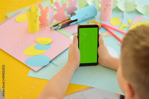 a close-up of a schoolboy who holds a mobile in his hands mobile phone with green screen. Easter crafts, craft tools and materials on a table. Festive spring crafts concept