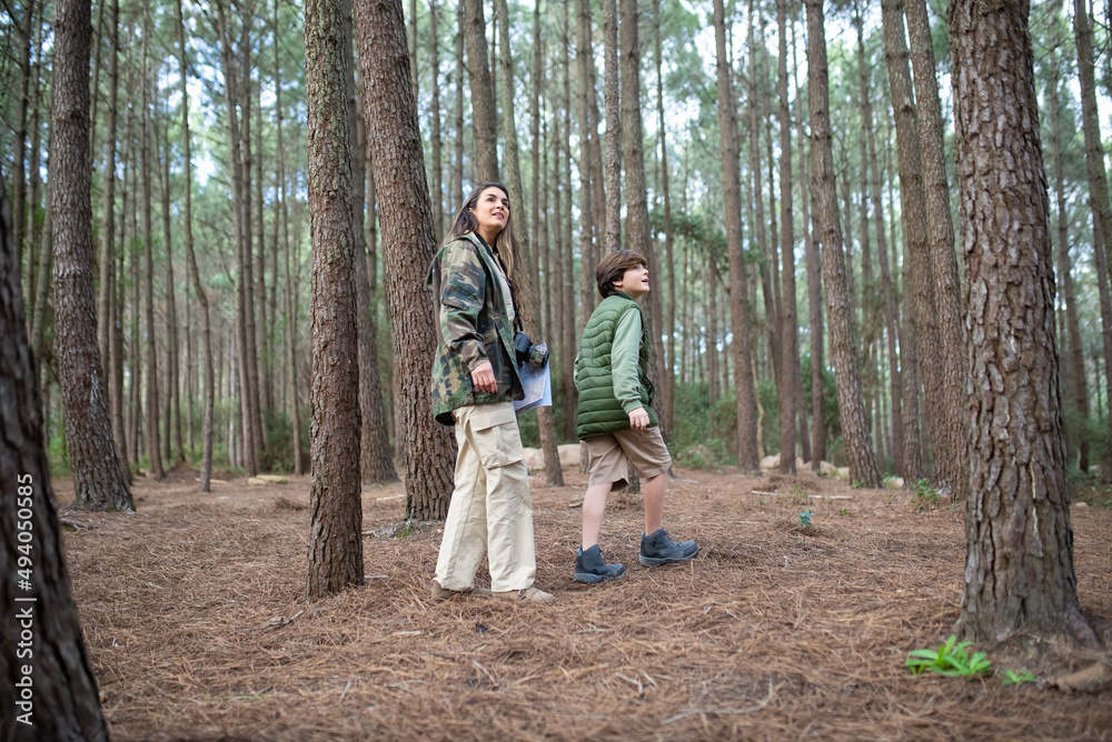 Content boy and woman with cameras walking in forest. Dark-haired mother and son in coats looking around. Parenting, family, leisure concept Content boy and woman with cameras walking in forest. Dark-haired mother and son in coats looking around. Parenting, family, leisure concept