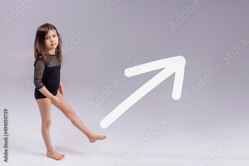 Strong little toddler girl raises her leg on a gray background. White arrow For women's empowerment, female power and feminism, sports, education and creative ideas of the future. Copy and space.